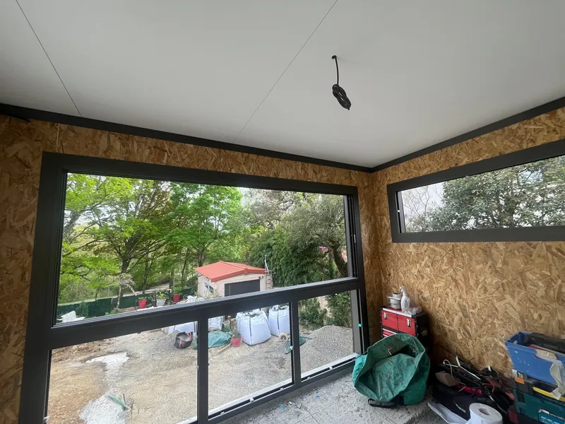 Pièce intérieure avec grandes fenêtres et cadres aluminium foncés donnant vue sur jardin extérieur